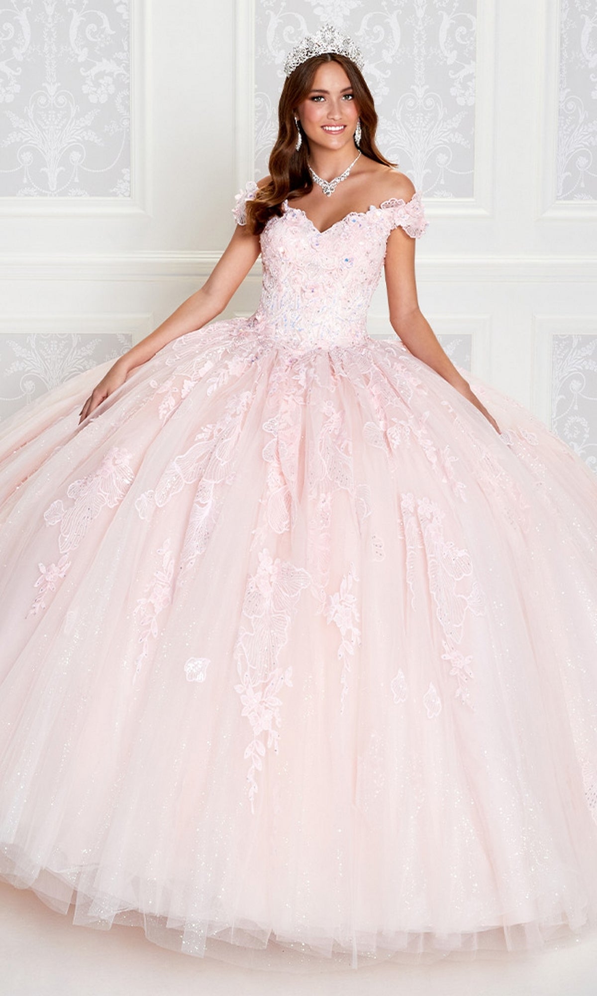 A young woman models the Princesa by Ariana Vara Pastel Quince Dress PR12270, a light pink off-the-shoulder ball gown with lace and sparkling details, posing indoors against a white decorative backdrop.